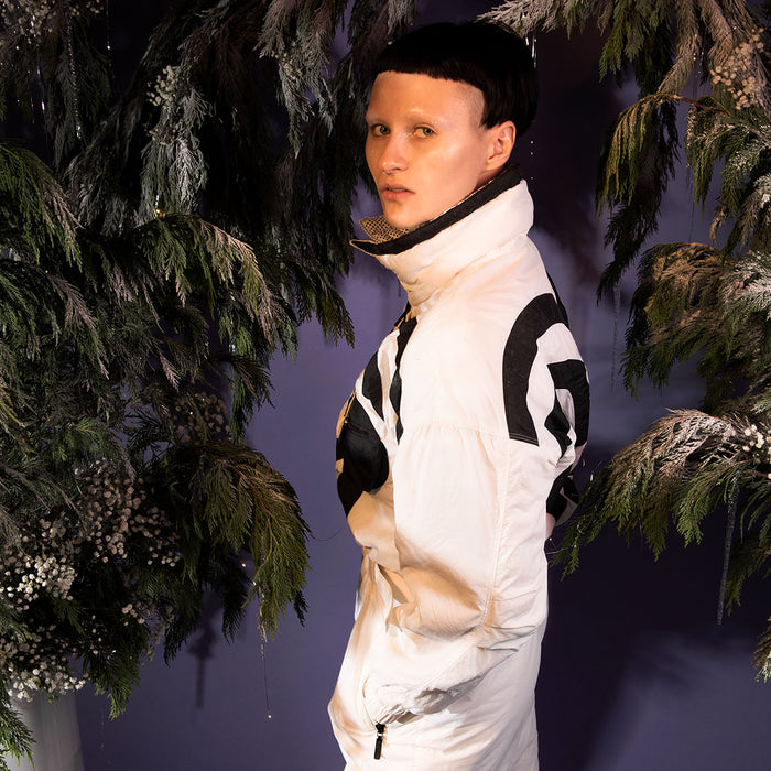 profile of model in vintage ski suit amongst pine installation. 