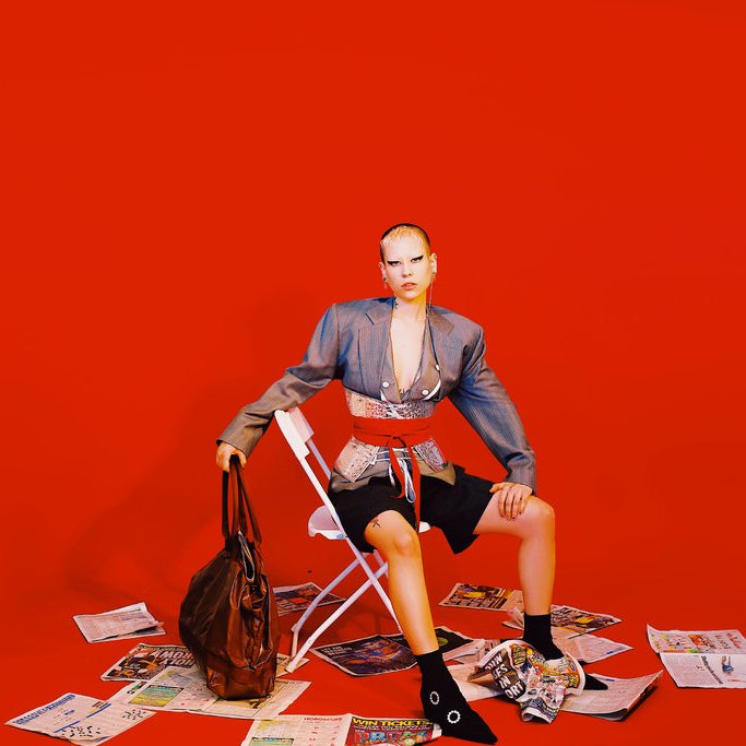 model seated on chair surrounded by news paper and holding a bag.