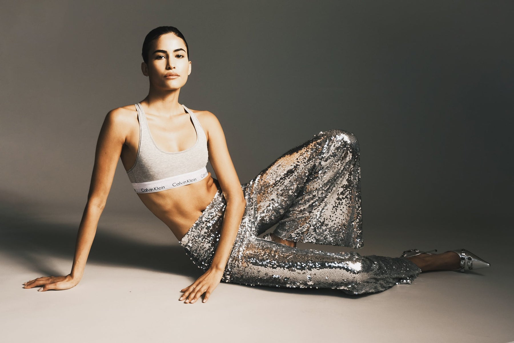 Woman in a Calvin Kline sports bra and sequined leggings seated on the floor with one knee pointed upward.