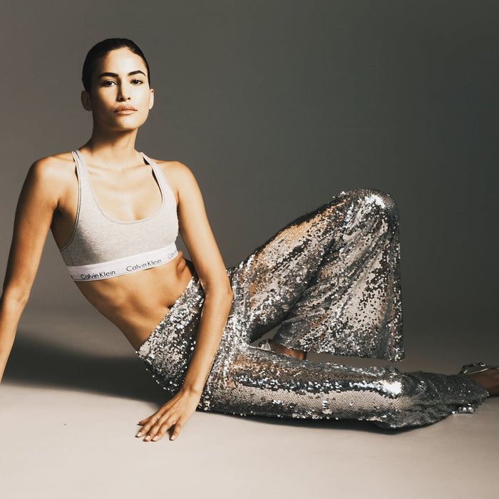 Woman in a Calvin Kline sports bra and sequined leggings seated on the floor with one knee pointed upward.