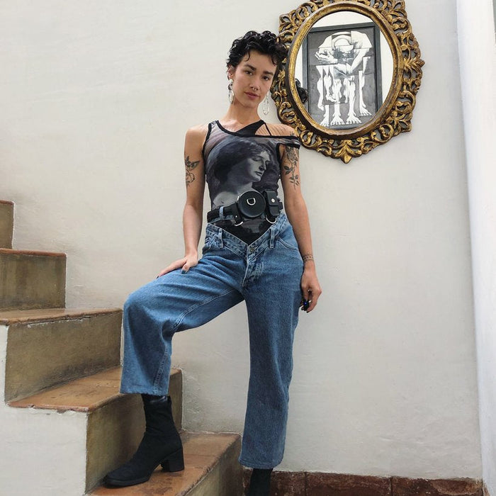 laura posing with their right leg on stairs, their right hand resting on the leg. behind them, a round mirror reflects a painting.