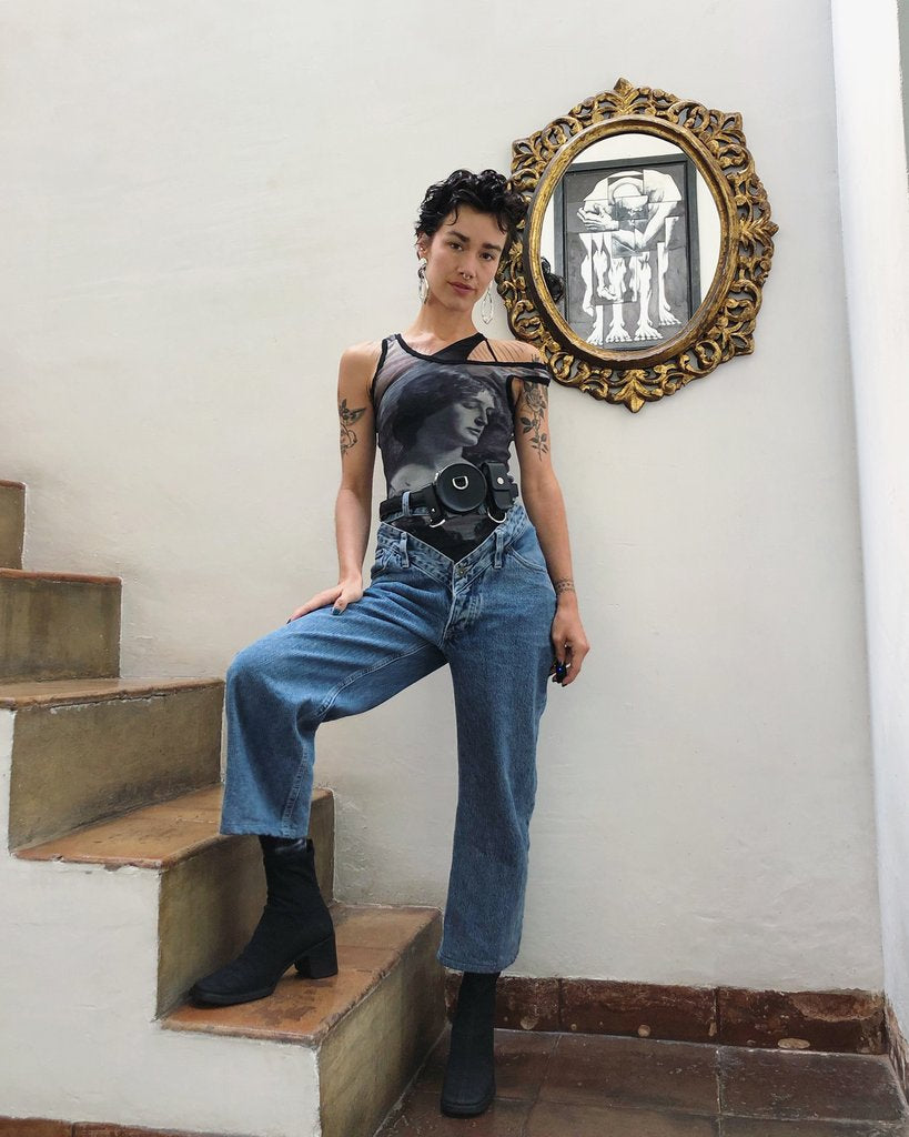 laura posing with their right leg on stairs, their right hand resting on the leg. behind them, a round mirror reflects a painting.