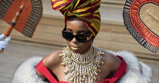 portrait of model seated with sunglasses head wrap and surrounded by fans.