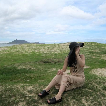 leah sitting in a field with a hat on, hand covering their face, with a mountain in the background.