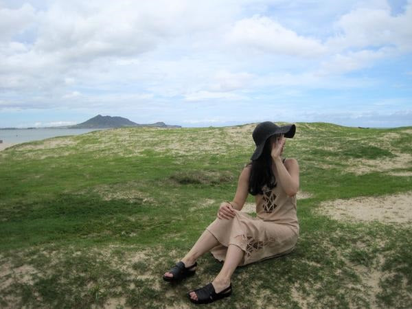 leah sitting in a field with a hat on, hand covering their face, with a mountain in the background.