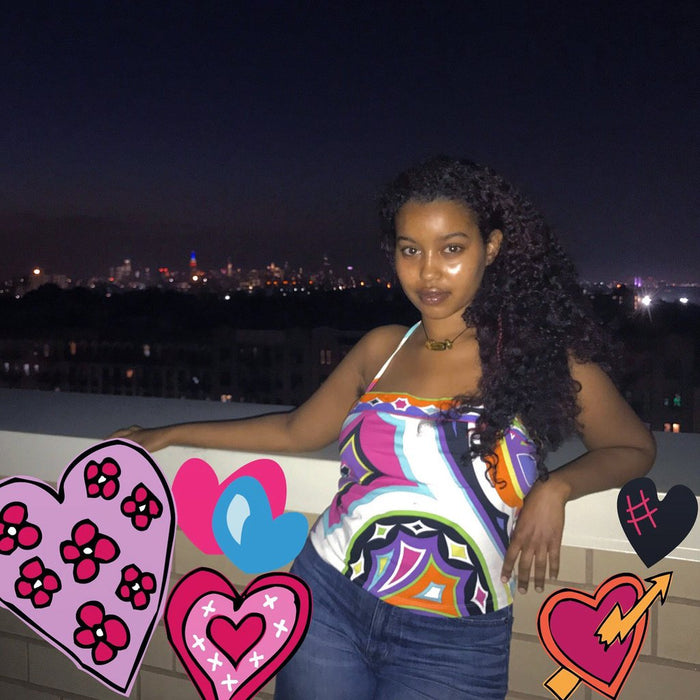 wilhelmina in printed tank and jeans leaning on brick barrier with night cityscape in background and heart graphics.