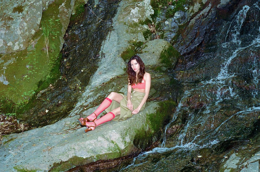model seated on rocks next to waterfall. 