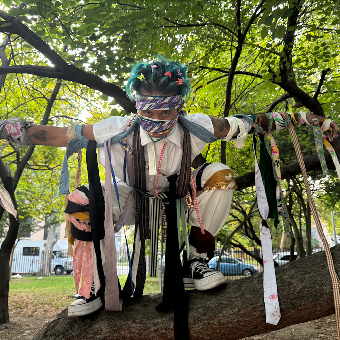 man posing like scarecrow in tree wearing avante garde ties and belts wrapped around limbs