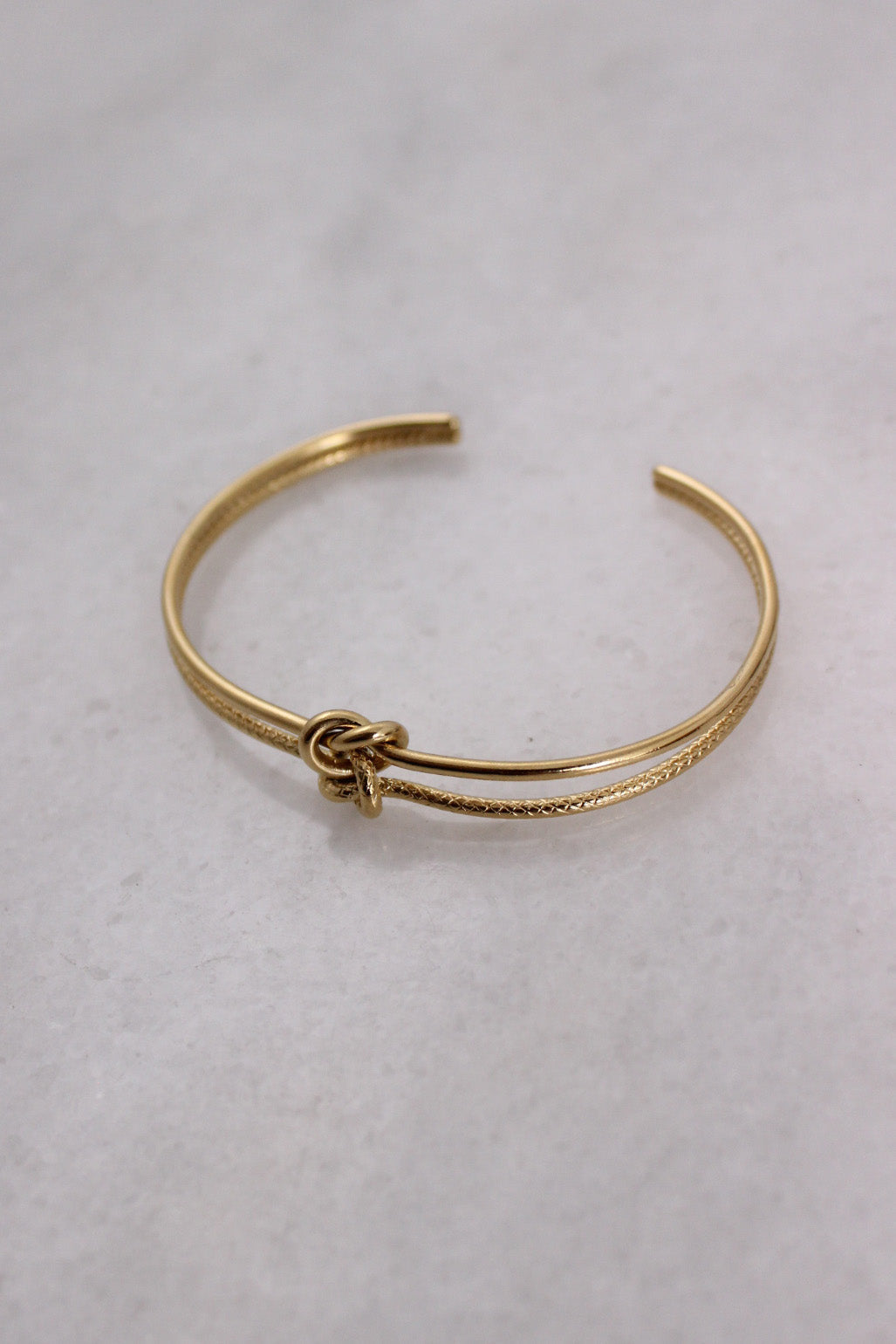 three-quarter front angle of gold-toned metal bracelet featuring smooth/textured base and knotted embellishment at center.