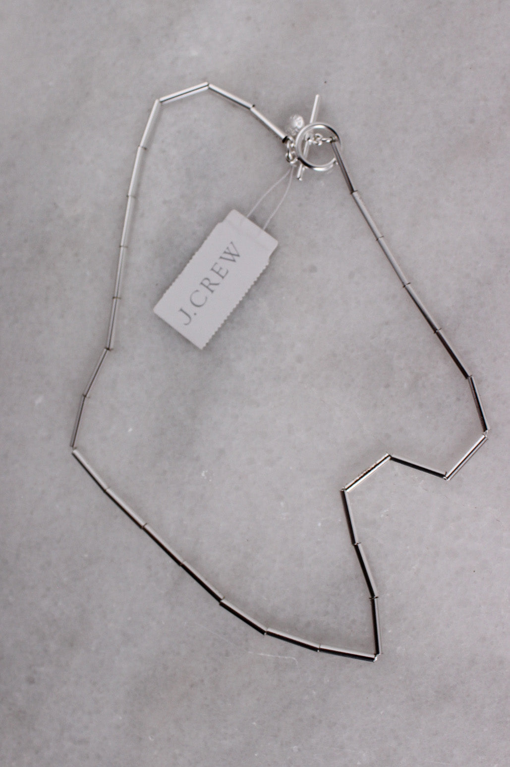 elevated angle j.crew silver toned metal necklace featuring cylindrical bead design, circular branded charm, and toggle closure laid on marble background.