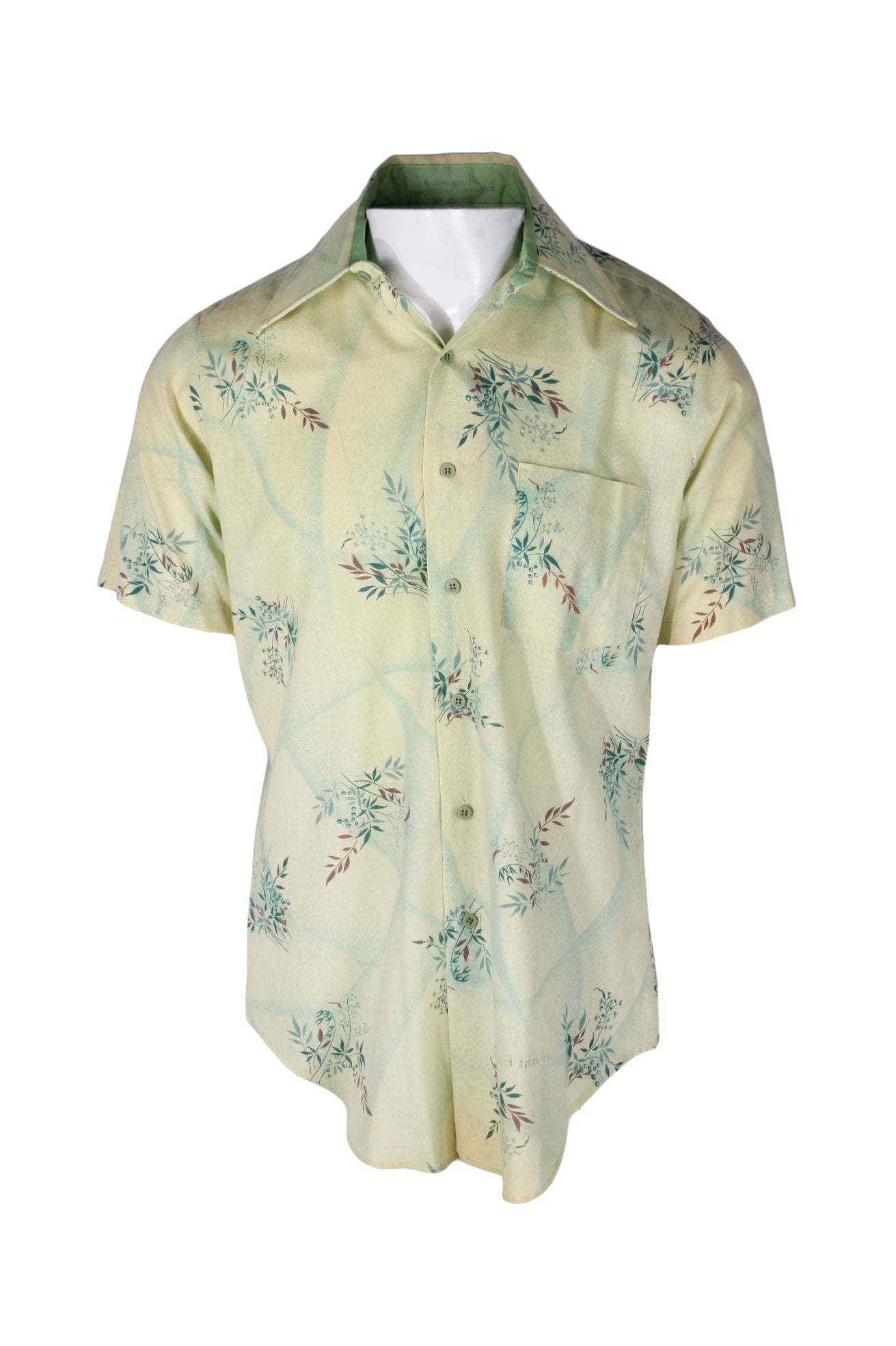 front angle vintage pale yellow/multicolor longline short sleeve button-up shirt on masculine mannequin torso featuring botanical/subtle geometric dotted print throughout, chest patch pocket, exaggerated pointed collar with built-in stays, and curved hemline.