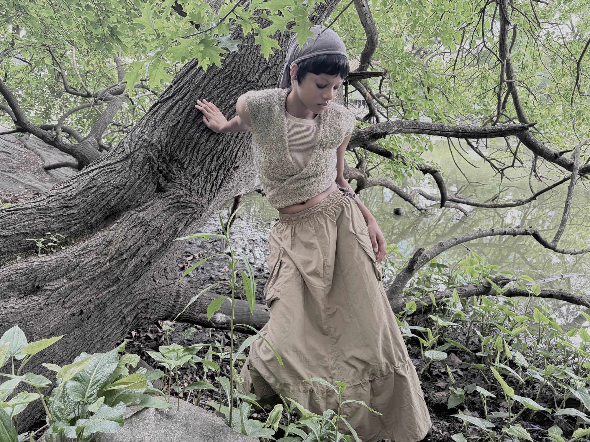 Person in a beige outfit standing in a forest with large trees and greenery.