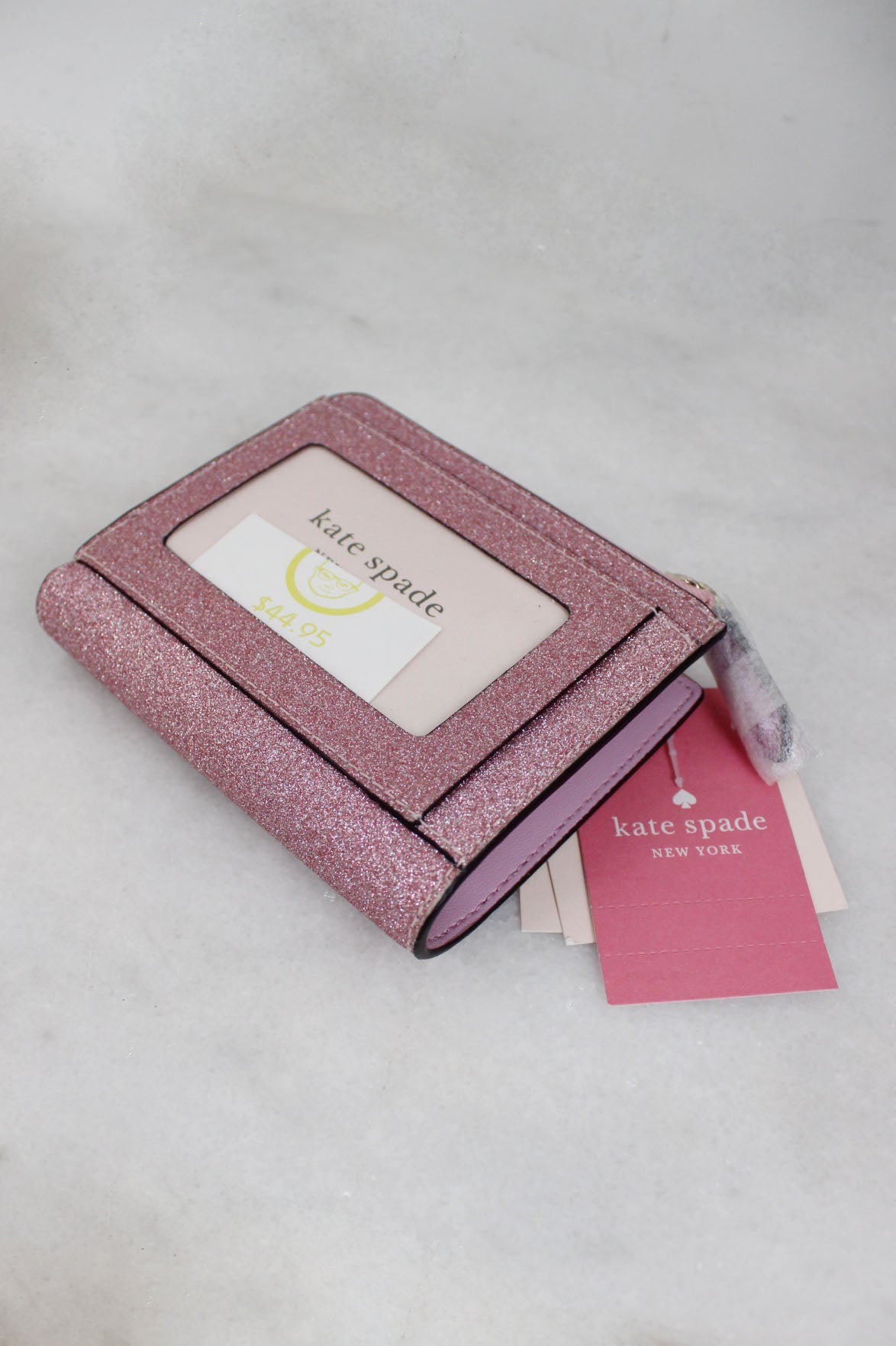 overhead view of kate spade pink ‘lola glitter’ small l-zip bifold wallet. features branding & l-zip compartment, interior 6-card slots, back clear window id compartment, metal pinmount with spade logo,  magnetic snap closure, bill fold, zip around coin compartment
