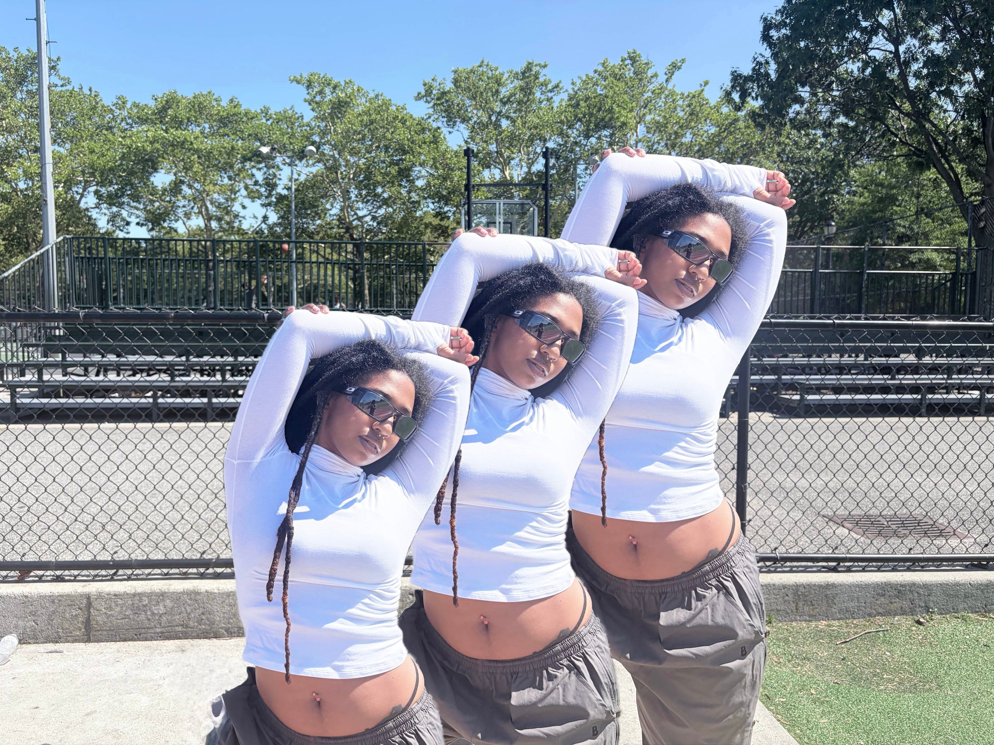 Three women posing outdoors in front of a chain-link fence and bleachers, wearing matching white long-sleeve crop tops and gray sweatpants.