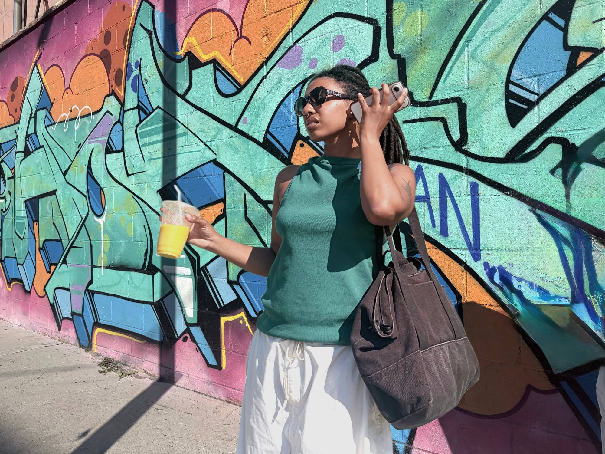 A woman wearing a green sleeveless top and white pants stands in front of a colorful graffiti wall, holding a drink and carrying a brown tote bag.