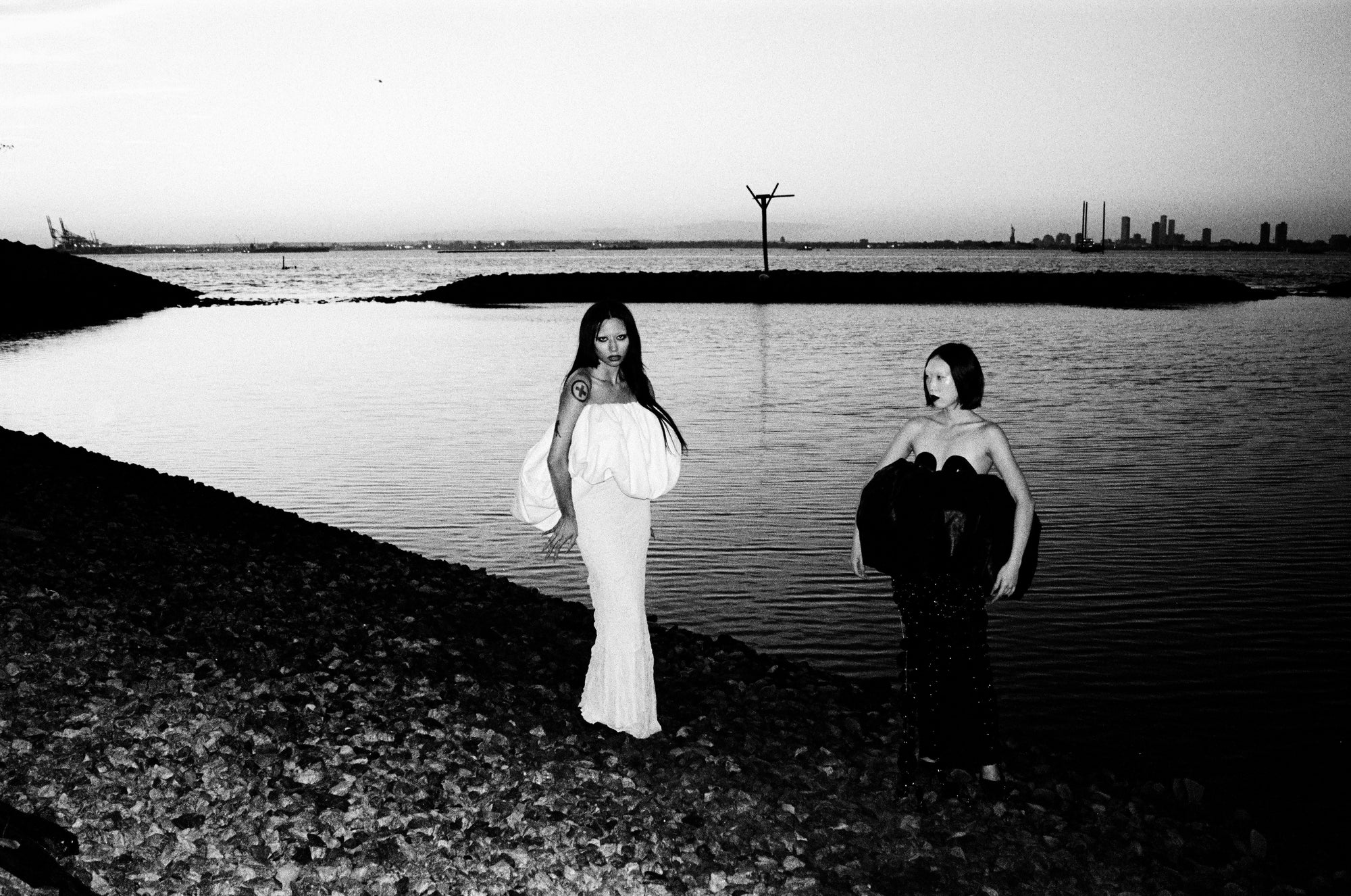 Two women standing by a body of water with a city skyline in the background.