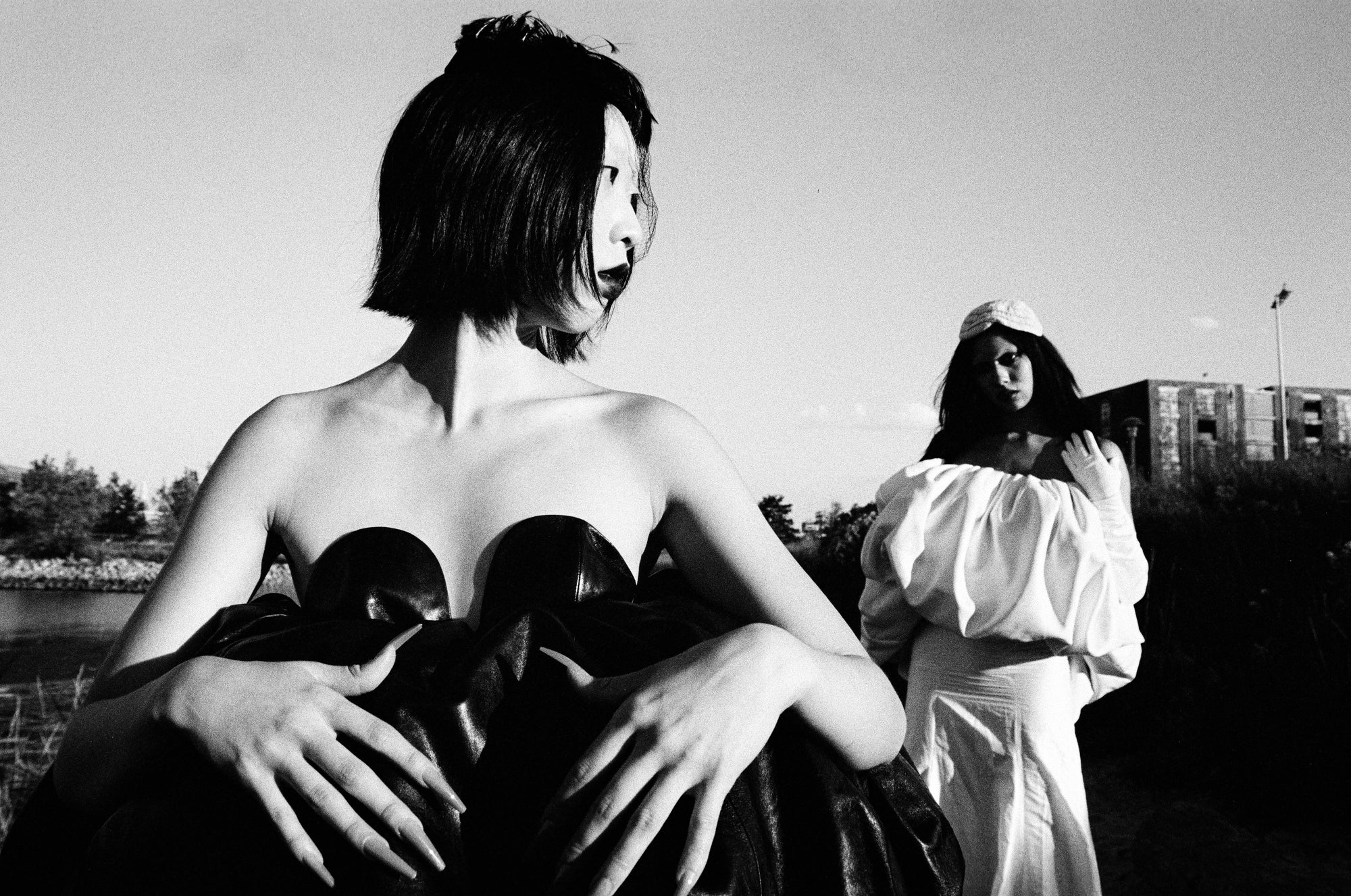 Black and white photograph of a person sitting with hands on knees, wearing a dress, with another person in a hooded outfit in the background.