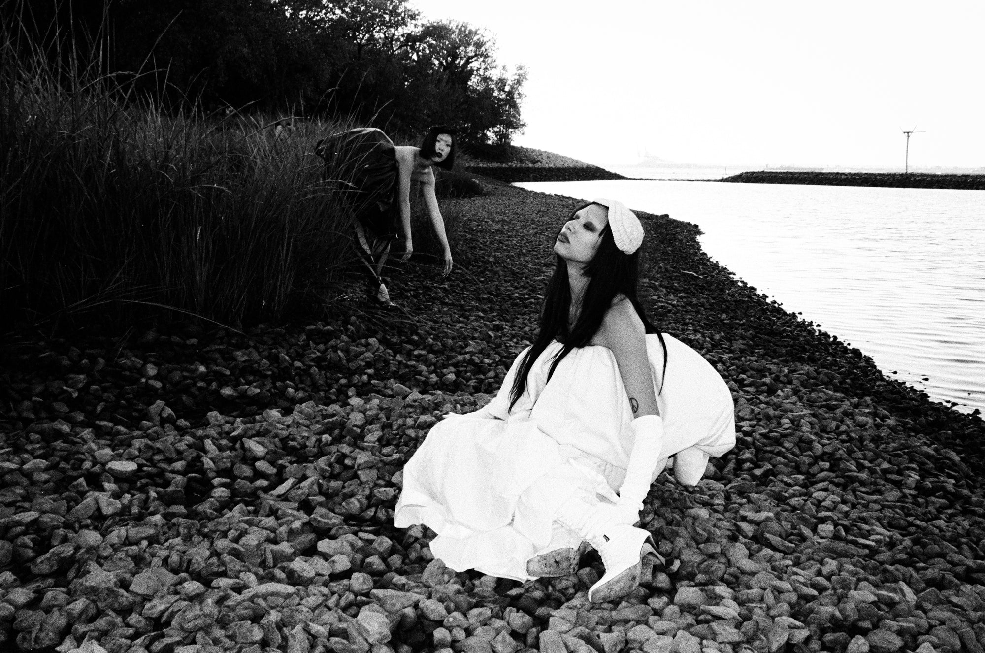 Woman in a white dress sitting on a rocky shore with water and grass in the background